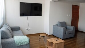 Ett vardagsrum med en grå soffa, en grå fåtölj, ett soffbord i trä och en platt-TV som är monterad på väggen.
