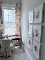 Una habitación con un ventanal grande, un escritorio con lámpara, una silla y varias fotografías enmarcadas de flores en la pared.