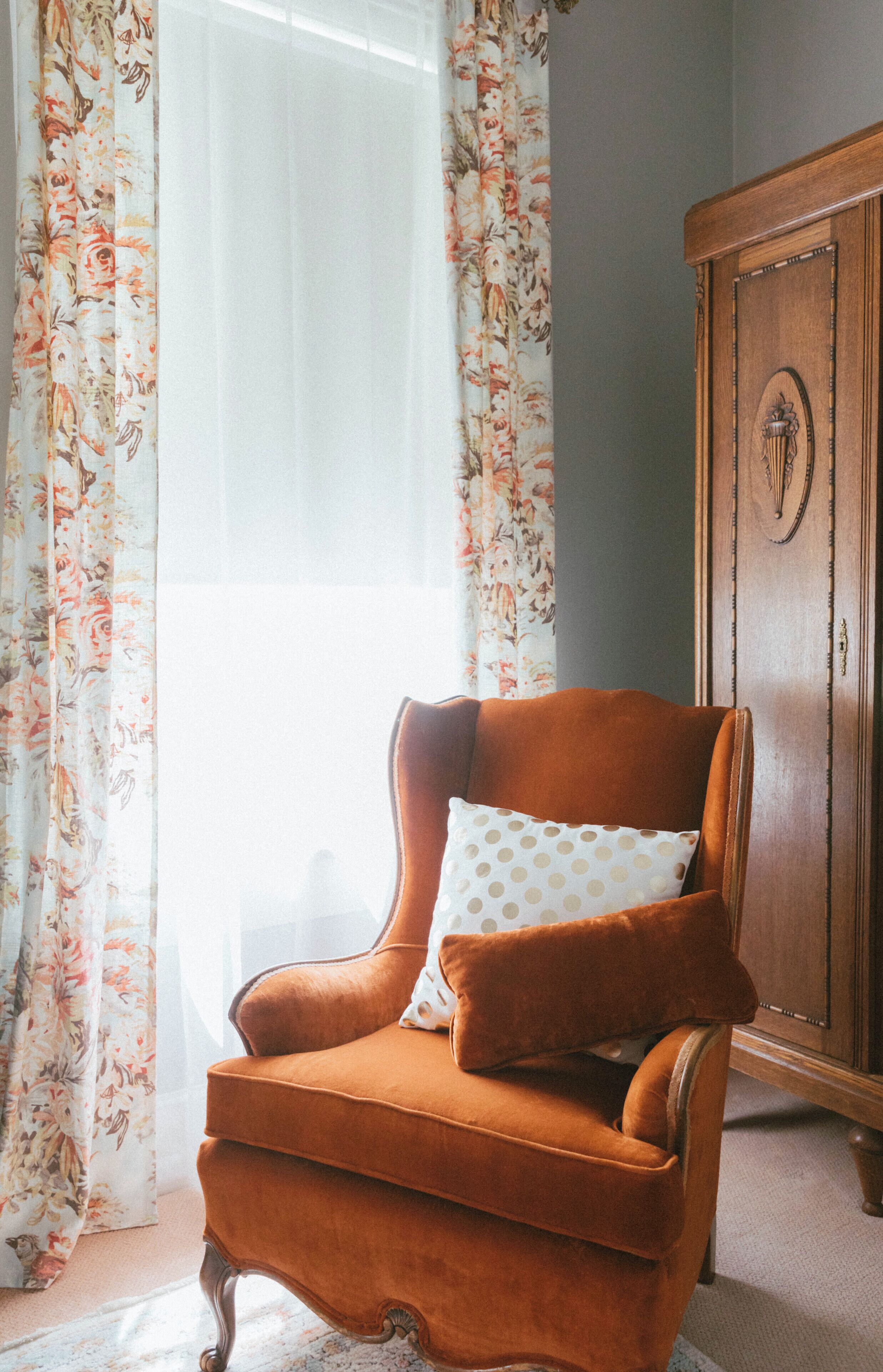 Habitación de esquina con un mueble de madera, un sillón tapizado marrón y un cojín estampado con lunares, además de cortinas florales.