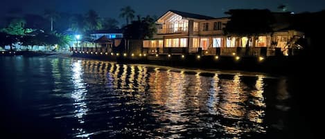 Una propiedad frente al agua con un edificio grande, un muelle y una masa de agua tranquila que refleja luces.