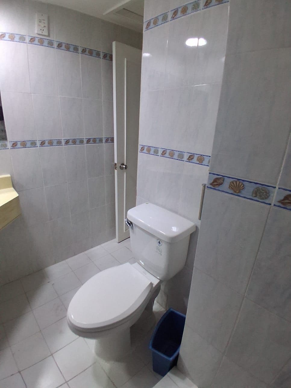 Un baño con inodoro, un cubo de basura azul y paredes con azulejos y un borde decorativo.
