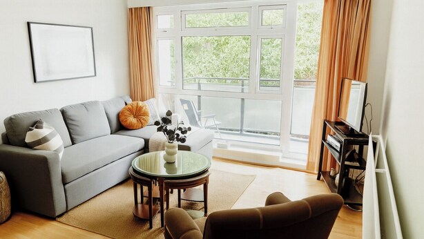 A living room with a grey sofa, a round glass table, a brown armchair, a TV stand, and a large window with curtains. - Bright and Cozy London Apartment (London)