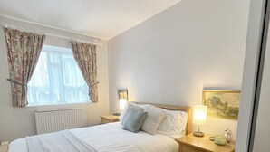 A bedroom with a bed, bedside lamps, a nightstand, a radiator, and a window with curtains. - Bright and Cozy London Apartment (London)