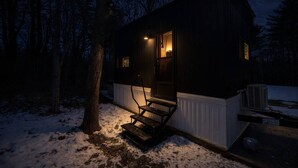 A cabin with a wooden exterior, a porch with a bench, and a lit interior visible through the door.