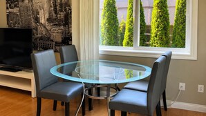 Un comedor con una mesa de vidrio rodeada por cuatro sillas, un televisor sobre un mueble bajo y una ventana con vistas a árboles.