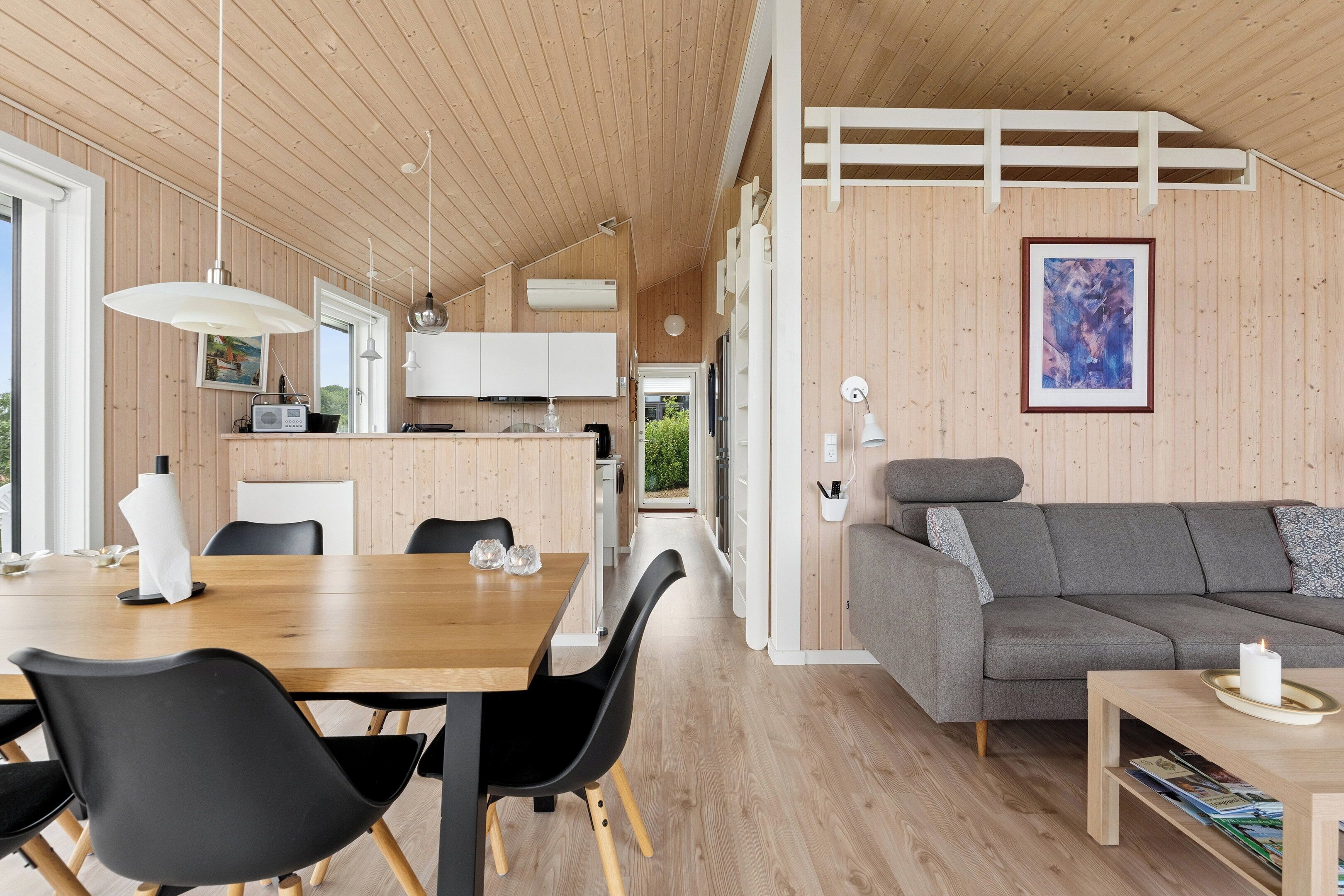 Un interior moderno con techo de madera, una zona de comedor con mesa y sillas, un sofá gris y un cuadro abstracto enmarcado en la pared.
