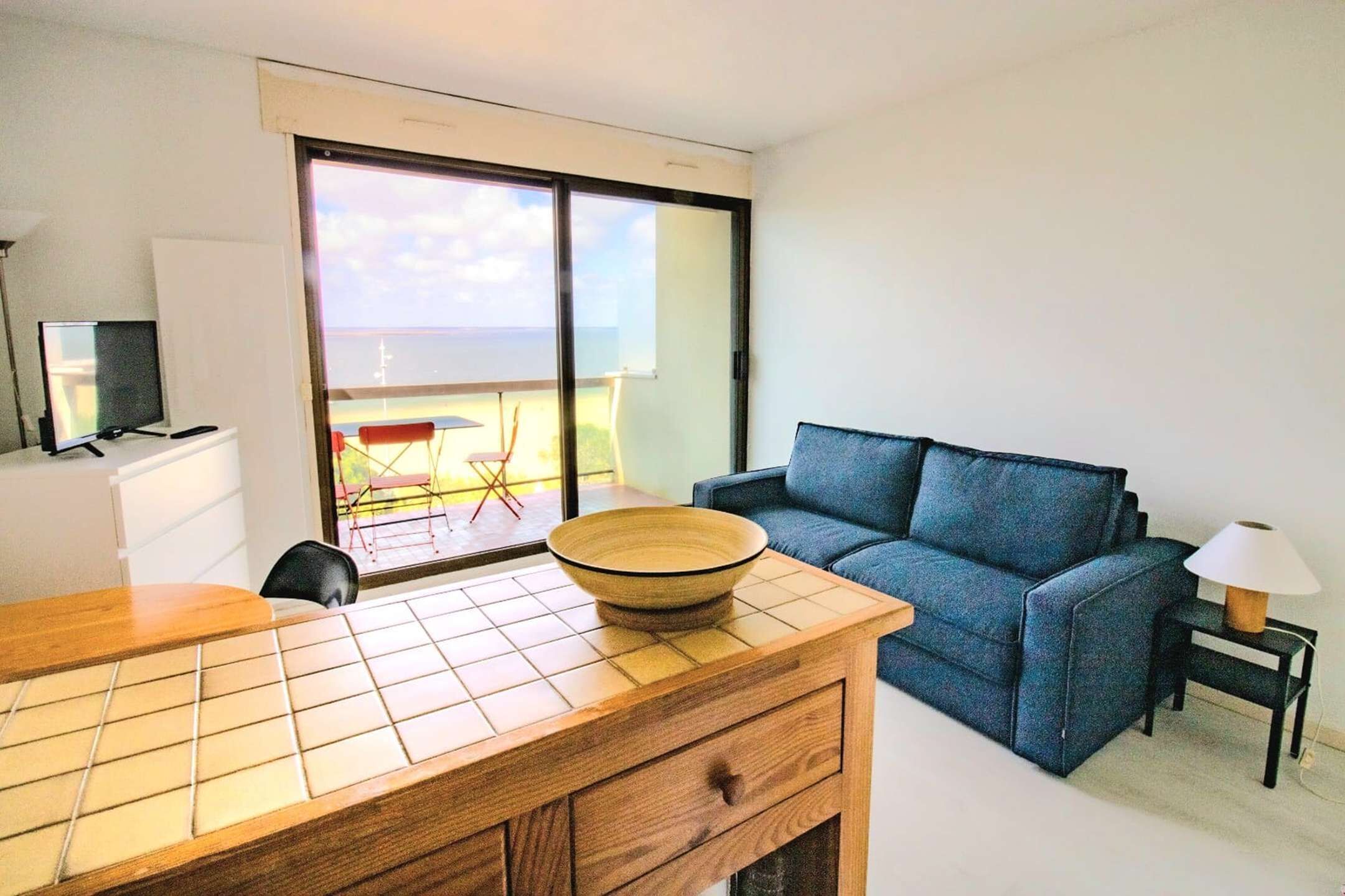 Una habitación moderna con una mesa de comedor de madera, un sofá azul, un televisor y un amplio ventanal con vistas a un balcón.
