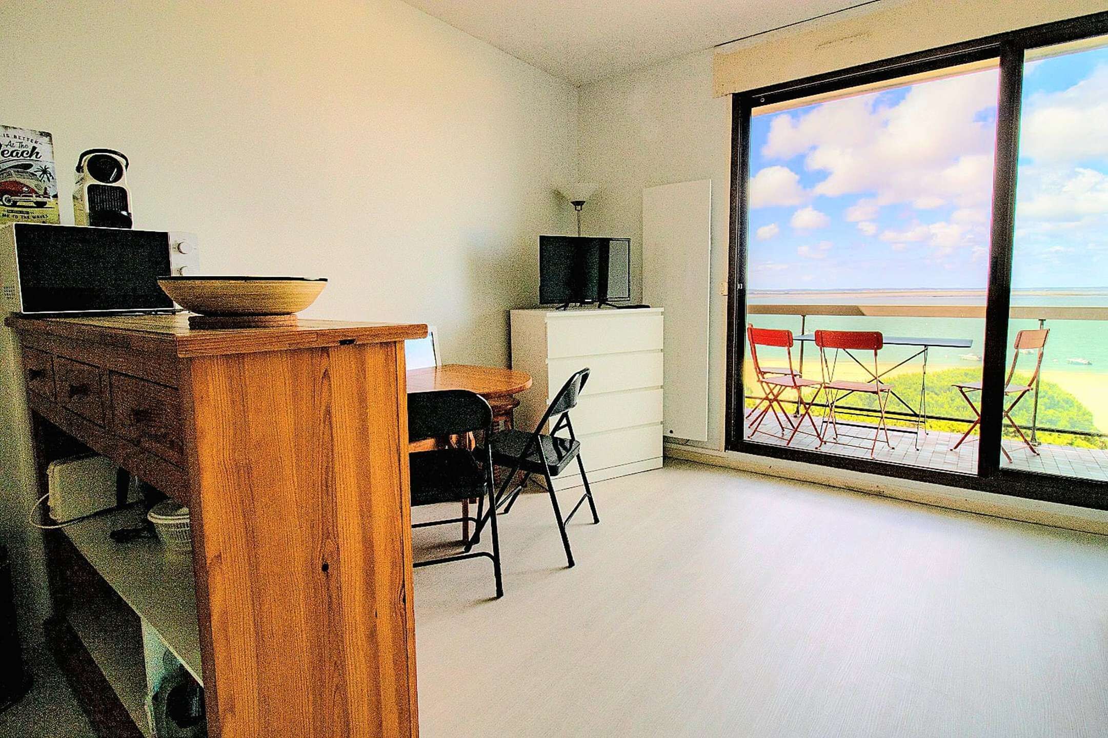 Ein Zimmer mit Schreibtisch aus Holz, Fernseher, Essbereich mit Stühlen und Blick auf den Strand durch große Fenster.