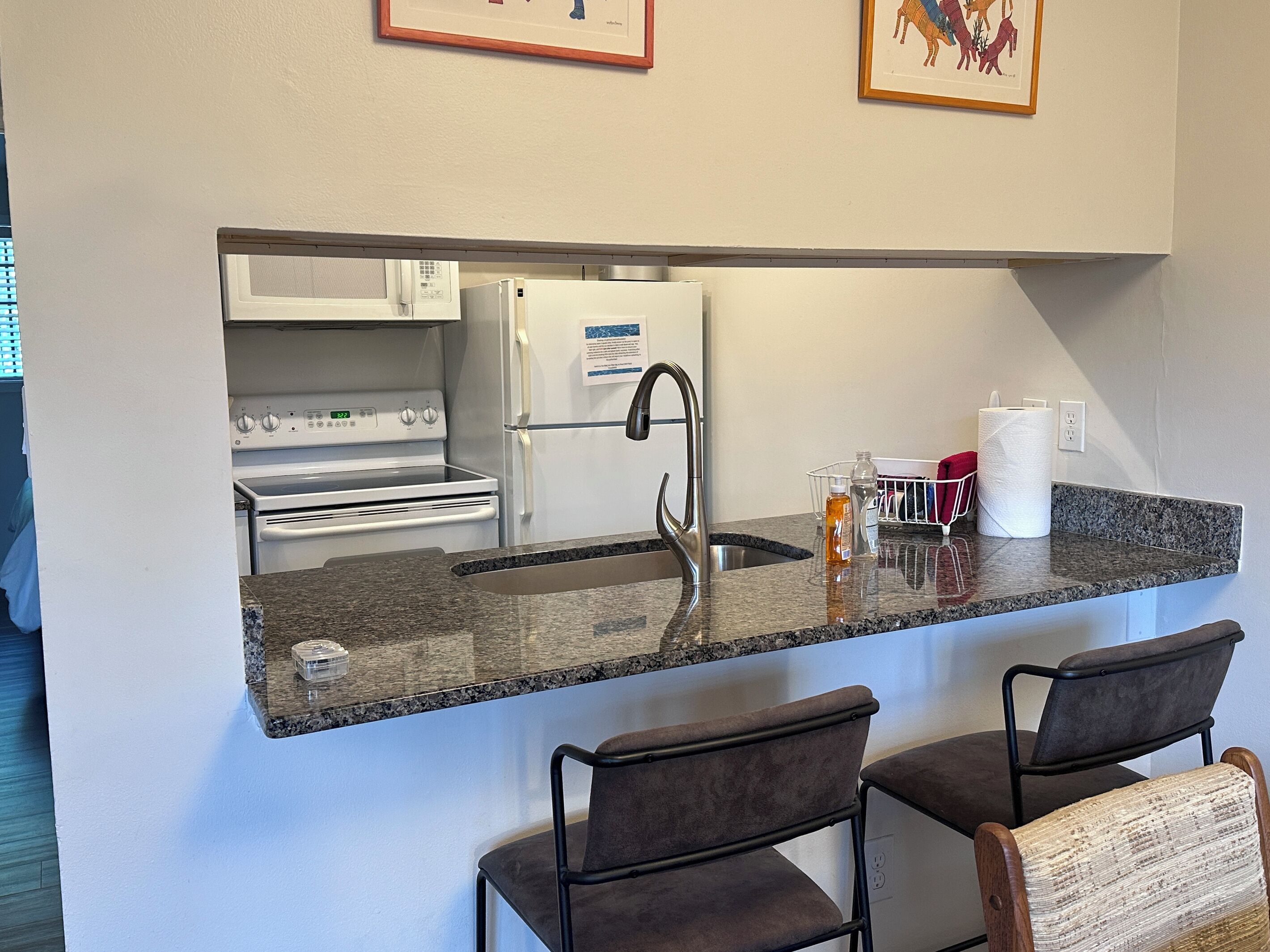 A compact kitchen with a built-in microwave, refrigerator, and stove. There are two bar stools and a small sink with a faucet.