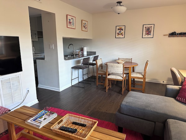 A living room with a TV, a dining area with chairs and a table, and a kitchen in the background.
