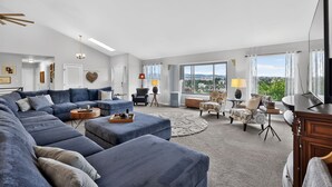 Ein geräumiges Wohnzimmer mit einer großen Eckcouch, einem runden Couchtisch und einem Fernseher. Es gibt verschiedene Sitzgelegenheiten, darunter ein Sessel und eine Zweisitzerliege. Große Fenster bieten einen Ausblick ins Freie.