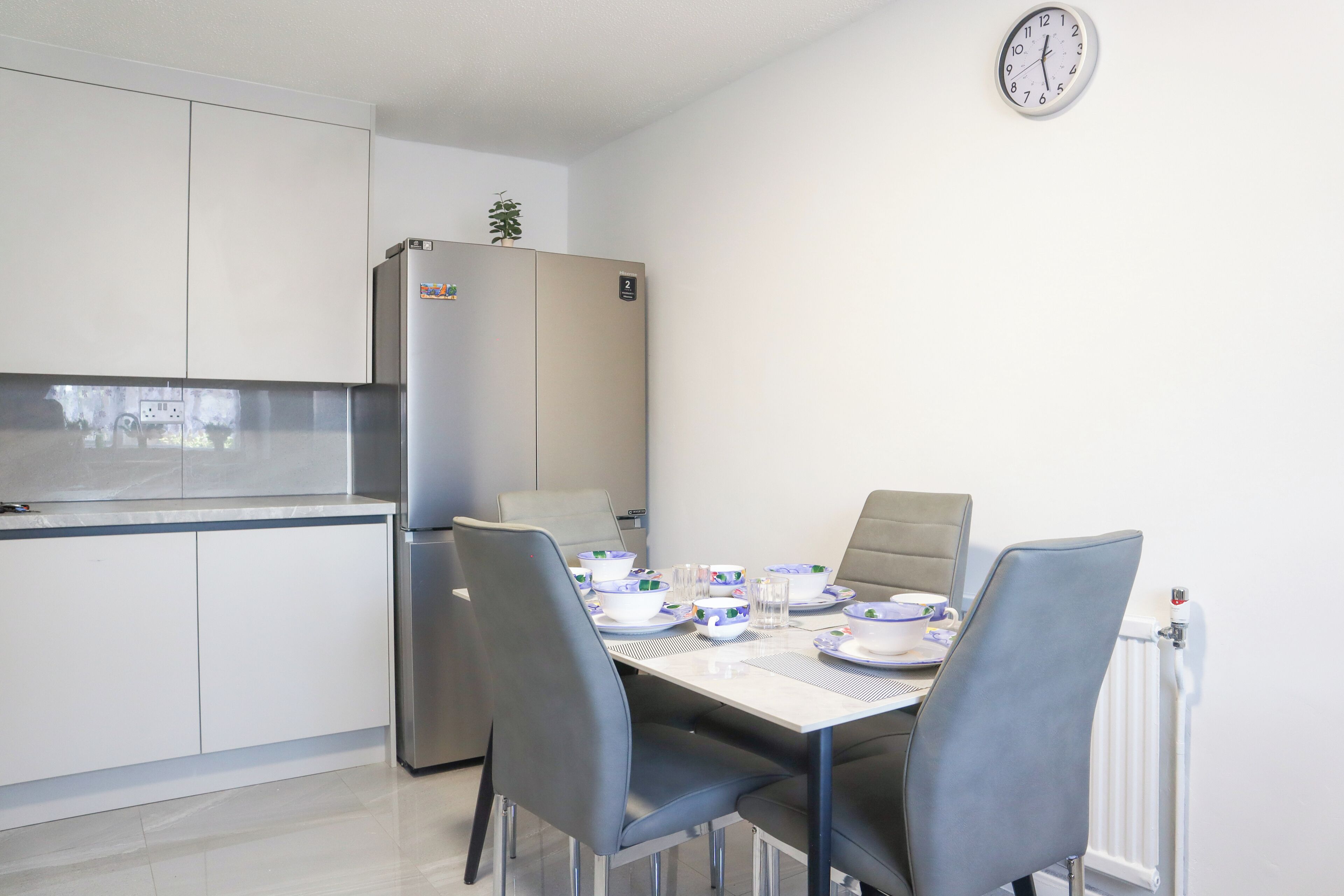 A modern kitchen with a dining table set for four, a refrigerator, and a clock on the wall.
