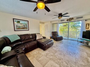 A living room with a brown leather sofa, a ceiling fan, a framed picture, a television, and a balcony with a view of palm trees. - Canal front. Ocean access via Tavernier Creek. New pool and hot tub. Sleeps 8 (Tavernier)