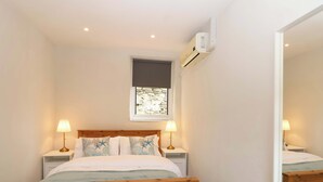A bedroom with a wooden bed, two bedside tables with lamps, a window with a view, and an air conditioning unit on the wall. - Walnut Major (Kingsbridge)