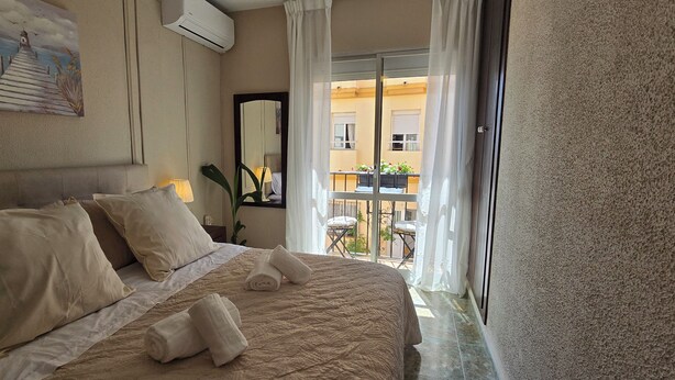 Une chambre d’hôtel avec un lit, des oreillers, un tableau, un climatiseur, un miroir et un balcon agrémenté de chaises et de plantes.