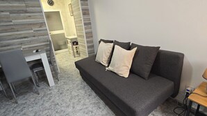 Une pièce moderne avec un canapé gris, deux coussins, une petite table avec une lampe et un bureau avec une chaise.