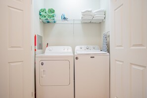 A laundry room with a front-loading washer and dryer, a shelf with folded towels, and a towel rack. - Spacious 3BR VistaCay Condo Near Disney, Universal & Orlando Attractions with Resort Amenities (Orlando)