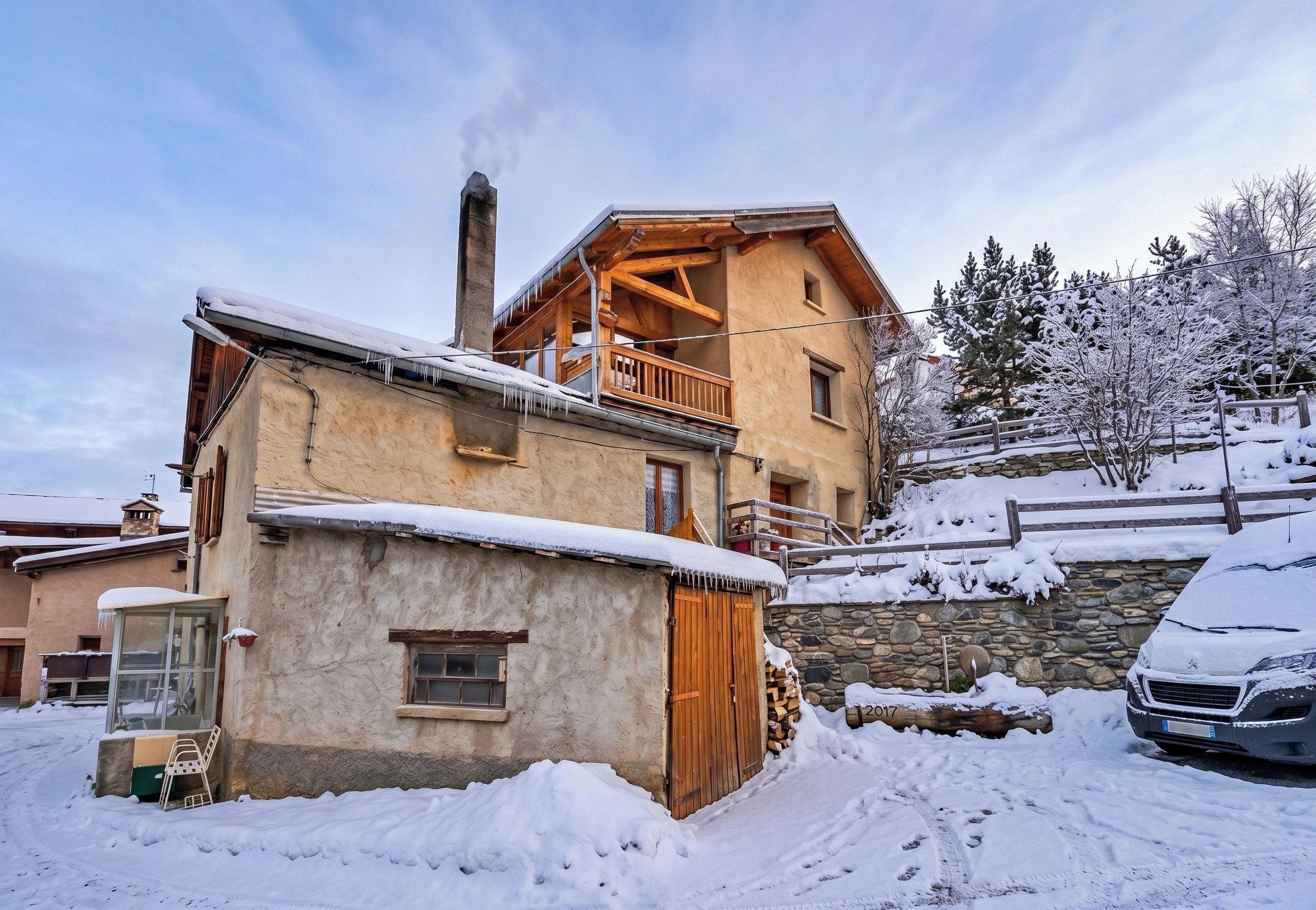 A two-story chalet with a balcony, a stone foundation, and a chimney emitting smoke.