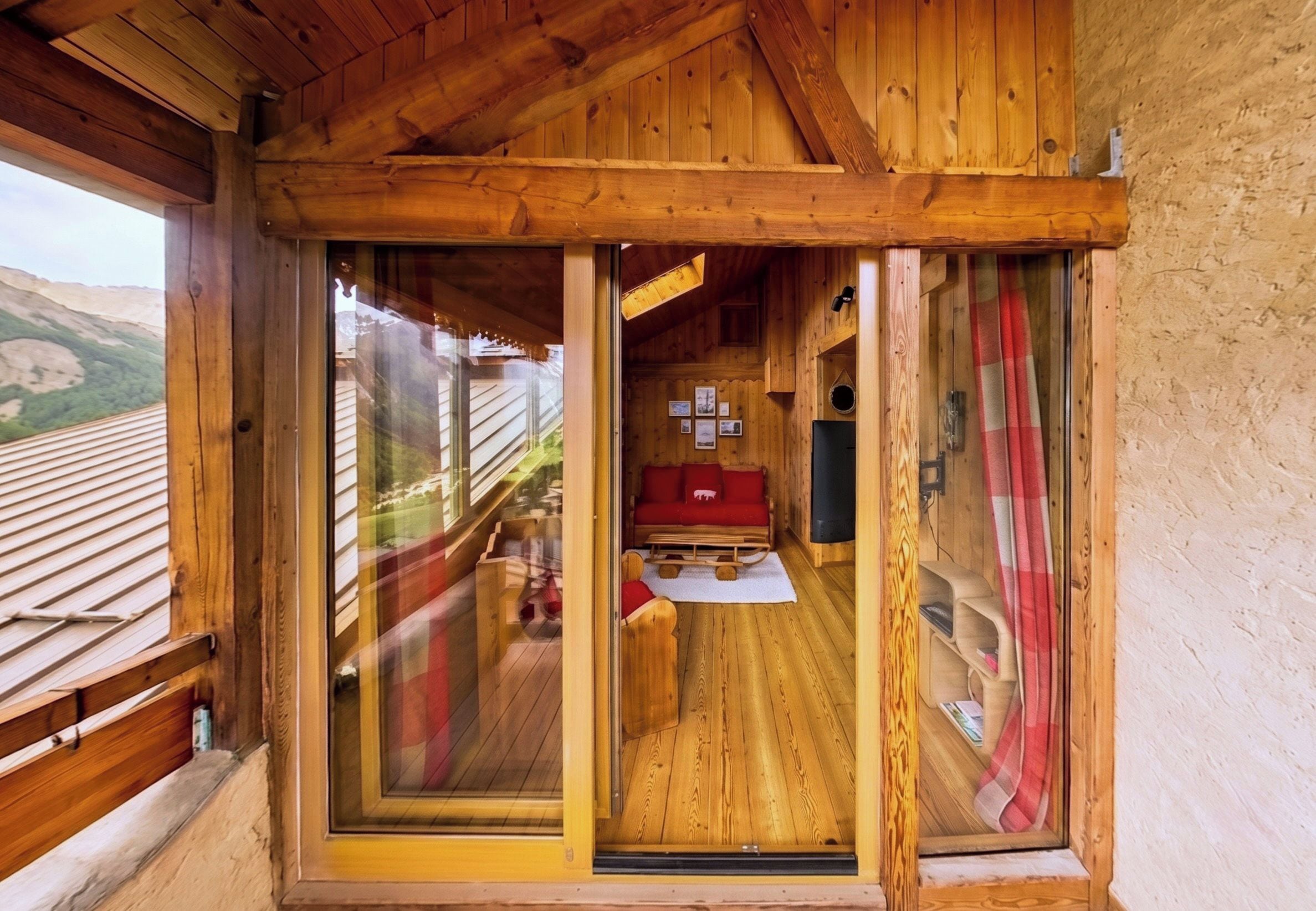 A wooden cabin with a sliding glass door leading to a cozy interior featuring a red sofa and a small table.