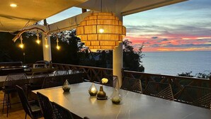 Een eethoek buiten met een tafel en stoelen, een hanglamp en uitzicht op zee bij zonsondergang.