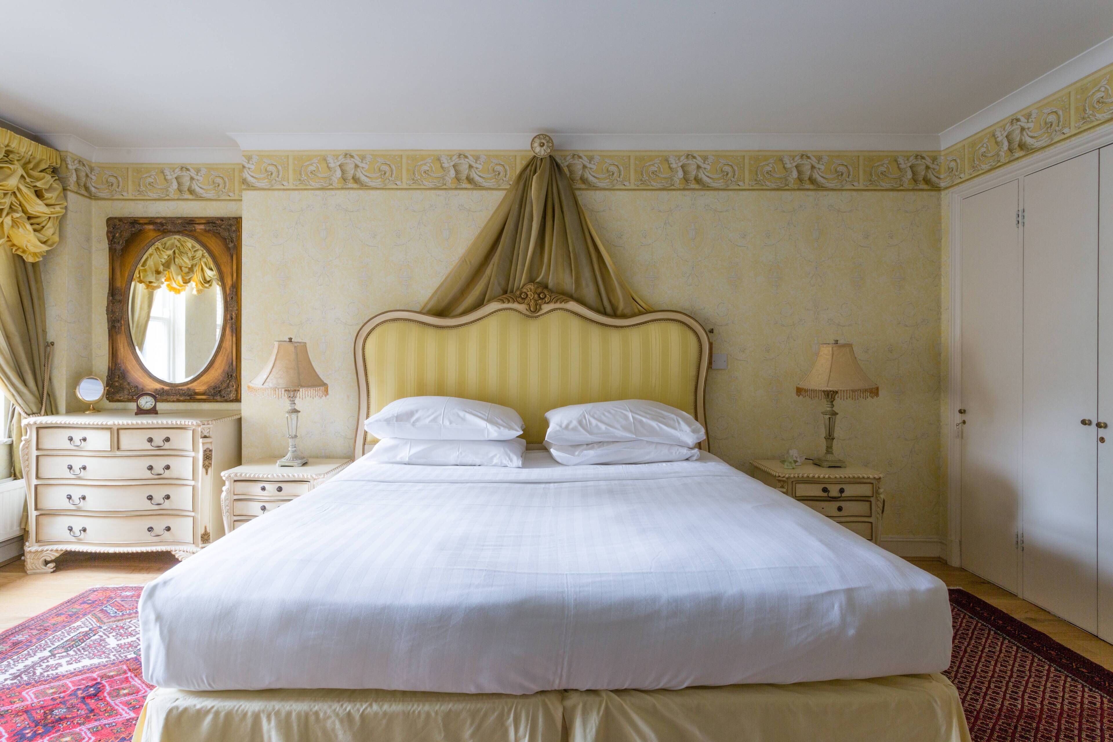 A large bed with a canopy, two bedside tables with lamps, a dresser with drawers, a mirror, and a wardrobe.