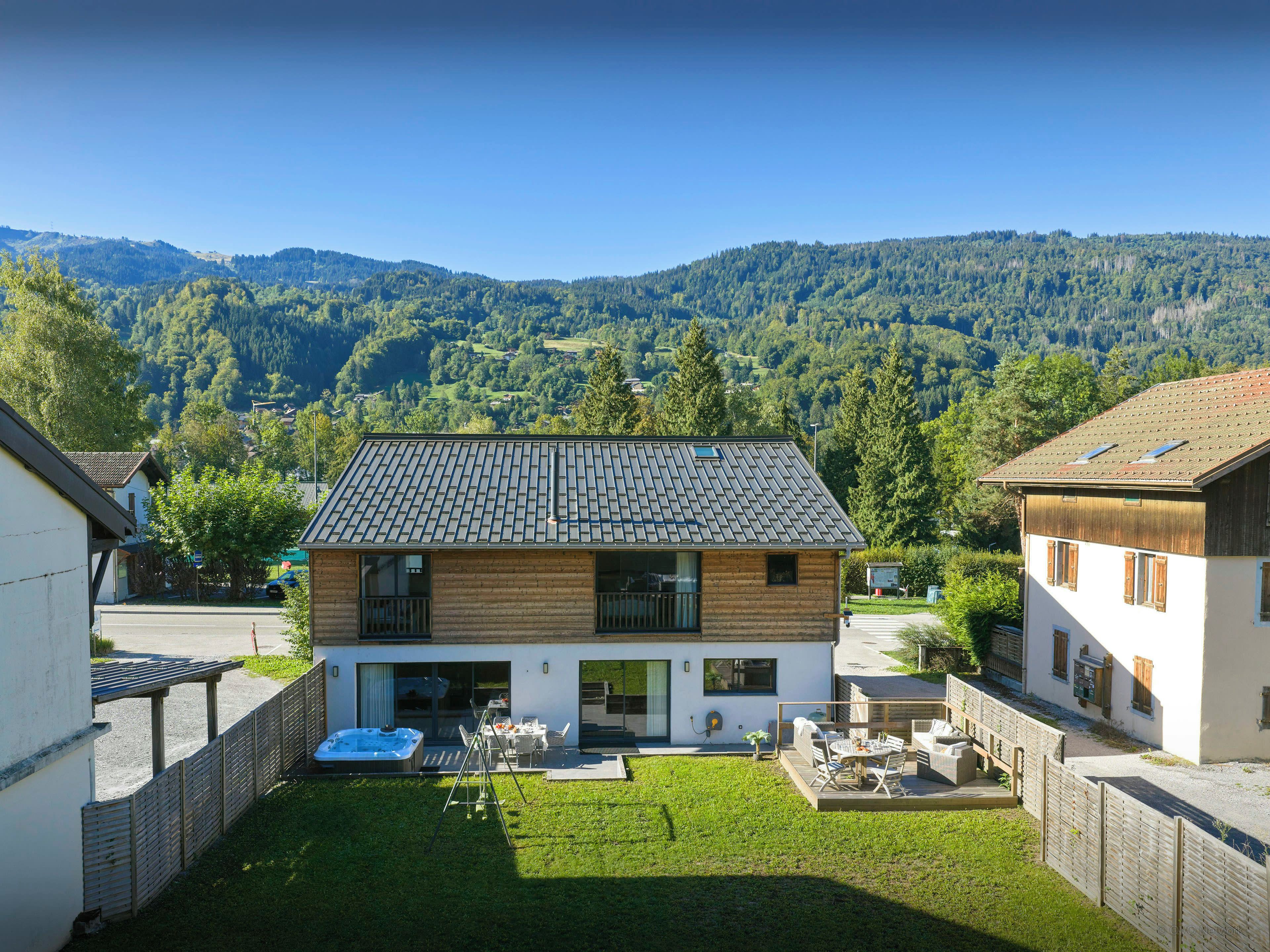 Una casa de dos pisos con techo inclinado, un patio con mobiliario exterior y vista a una cordillera boscosa.