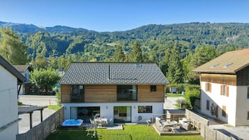 Una casa de dos pisos con techo inclinado, un patio con mobiliario exterior y vista a una cordillera boscosa.