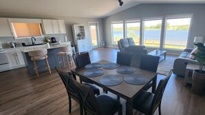 Een moderne keuken met witte kasten, een eethoek met een houten tafel en stoelen, en een groot raam met uitzicht op een waterpartij.