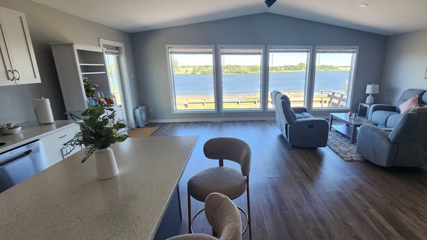 Een ruime woonkamer met een groot raam met uitzicht op een waterpartij, een keuken met moderne apparatuur en een zithoek met een grijze bank en fauteuils.