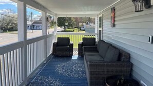 A covered porch with two wicker chairs and a sofa, a blue patterned rug, and a view of a residential street.