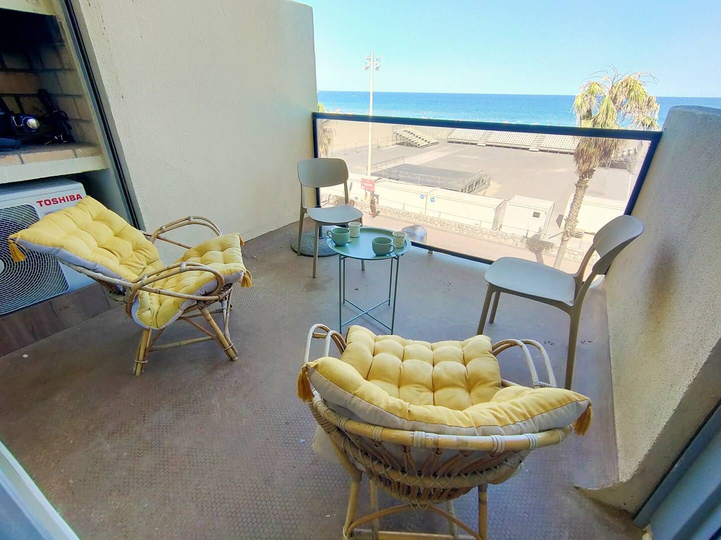A balcony with wicker chairs and a small table, overlooking a beach and ocean.