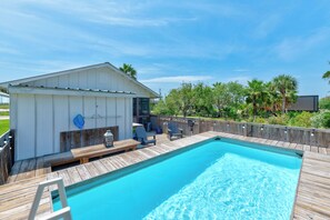 A swimming pool with a wooden deck and lounge chairs, a small shed, and a wooden fence with trees and a clear sky in the background.