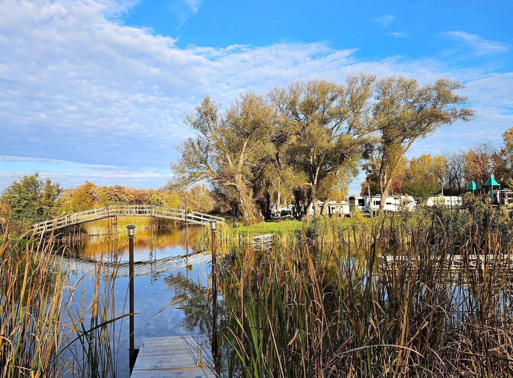 Escena serena junto al lago, con un puente de madera, agua tranquila y árboles al fondo.