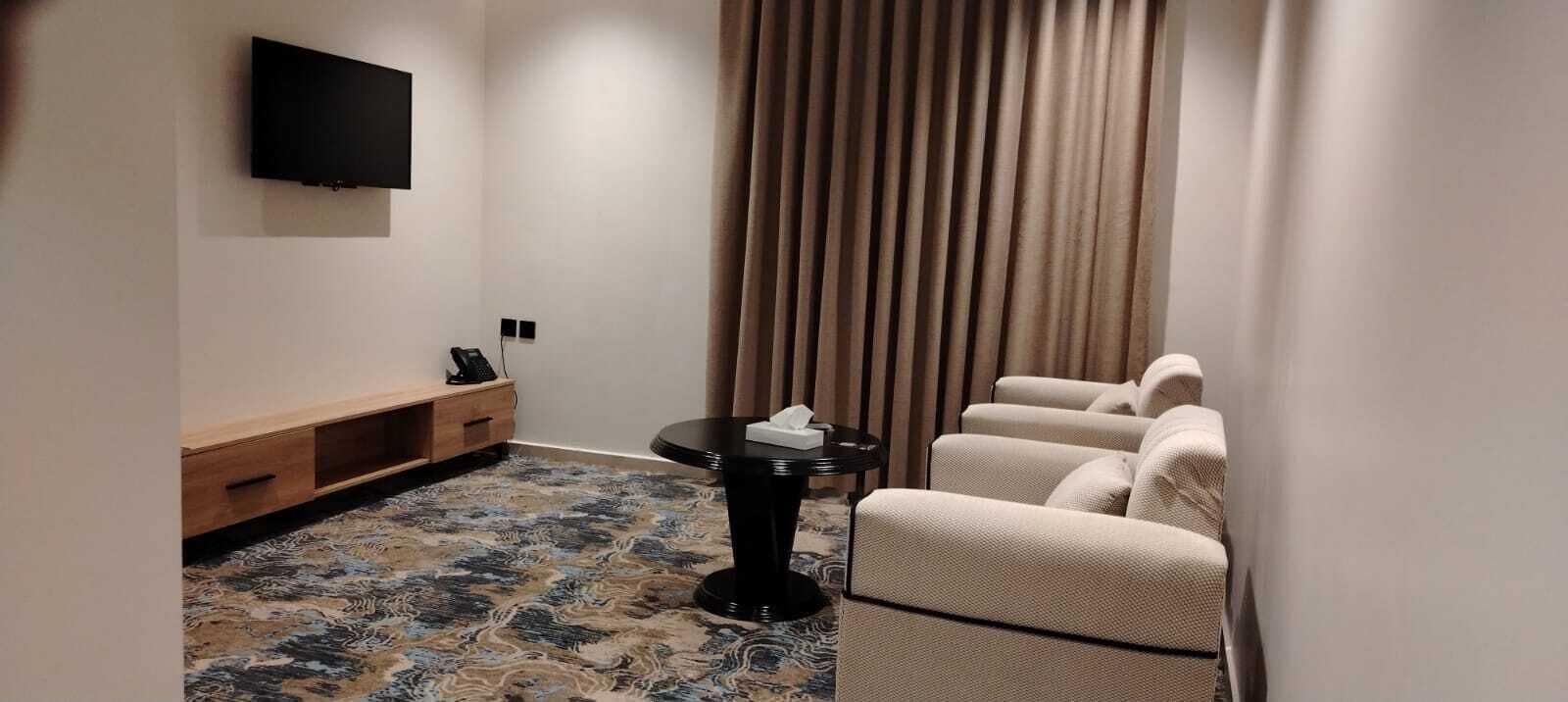 A hotel room with a sofa set, a small table with a telephone, a TV mounted on the wall, and a patterned carpet.