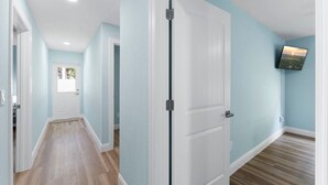A hallway with light blue walls, wooden flooring, a white door, and a mounted TV. - Simply Divine Beach Home | Walk to Beach | Firepit | Pets | Sleeps 8 (Palm Coast)