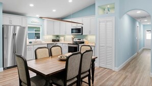 A modern kitchen with white cabinets, a stainless steel refrigerator, and a dining area with a wooden table and chairs. - Simply Divine Beach Home | Walk to Beach | Firepit | Pets | Sleeps 8 (Palm Coast)