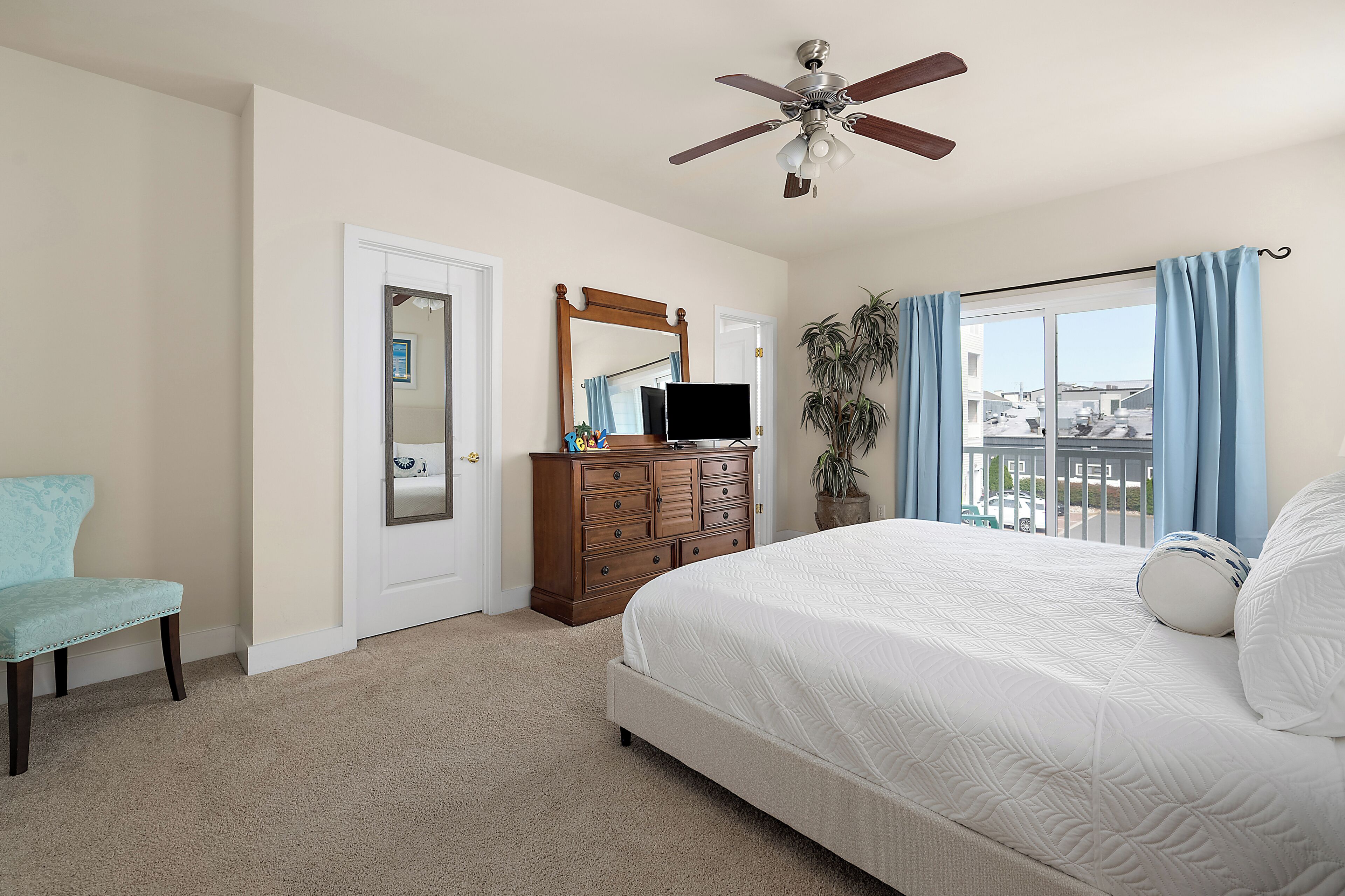 A bedroom with a bed, a dresser, a chair, a ceiling fan, a mirror, and a window with curtains.