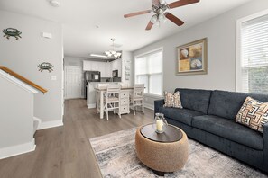 A modern living room with a dark grey sofa, a round woven coffee table, and a kitchen in the background. - Midtown 3BR Townhouse, Walk to Beach, Sleeps 10, Linens Included (Ocean City)