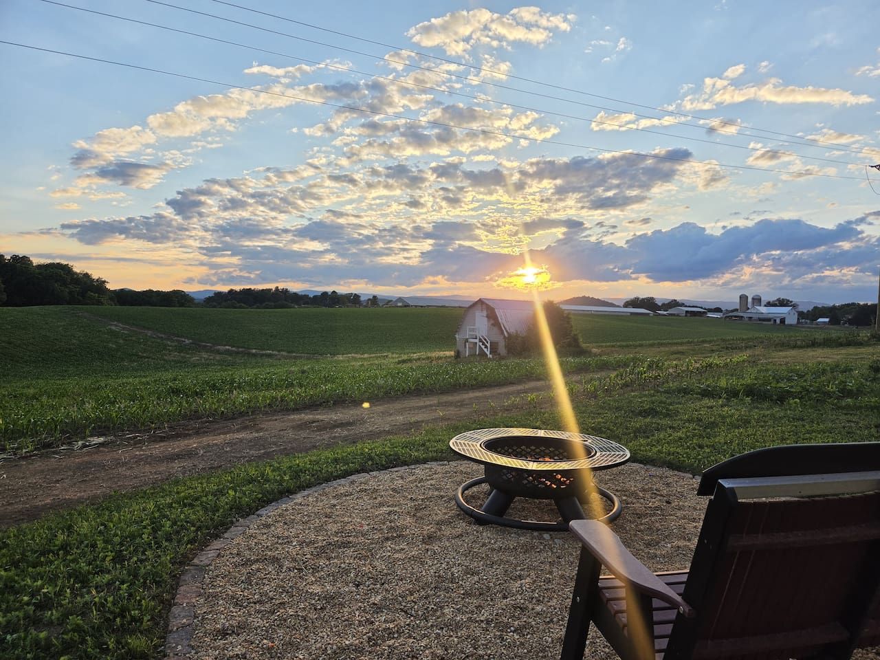 A gravel path leading to a circular outdoor seating area with a fire pit, surrounded by a grassy field and a barn in the background.