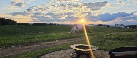 A gravel path leading to a circular outdoor seating area with a fire pit, surrounded by a grassy field and a barn in the background.