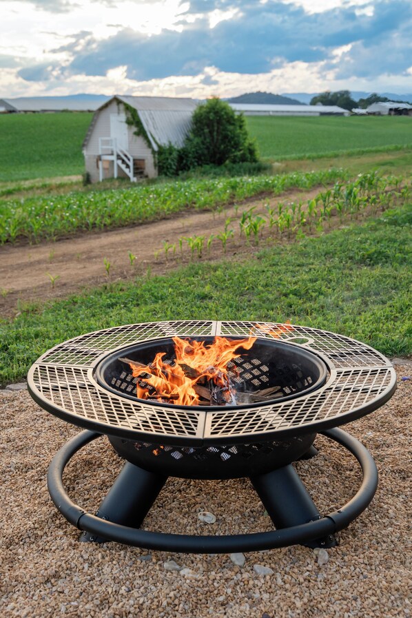 A circular outdoor fire pit with flames, set on a gravel surface with a rural landscape in the background.