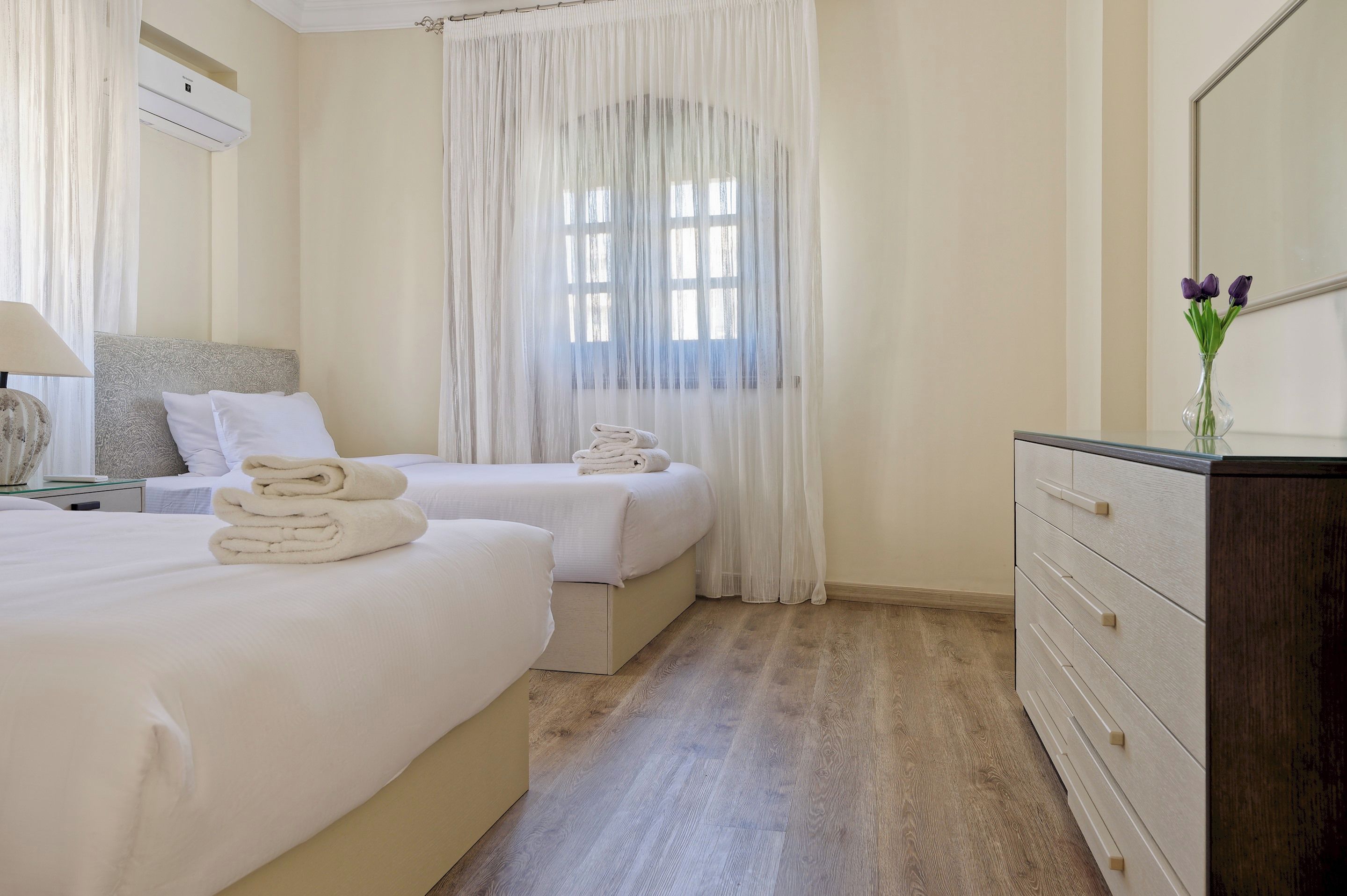 Habitación de hotel con dos camas, un aparador de madera, un espejo, una ventana con cortinas traslúcidas y una mesita de noche con lámpara.