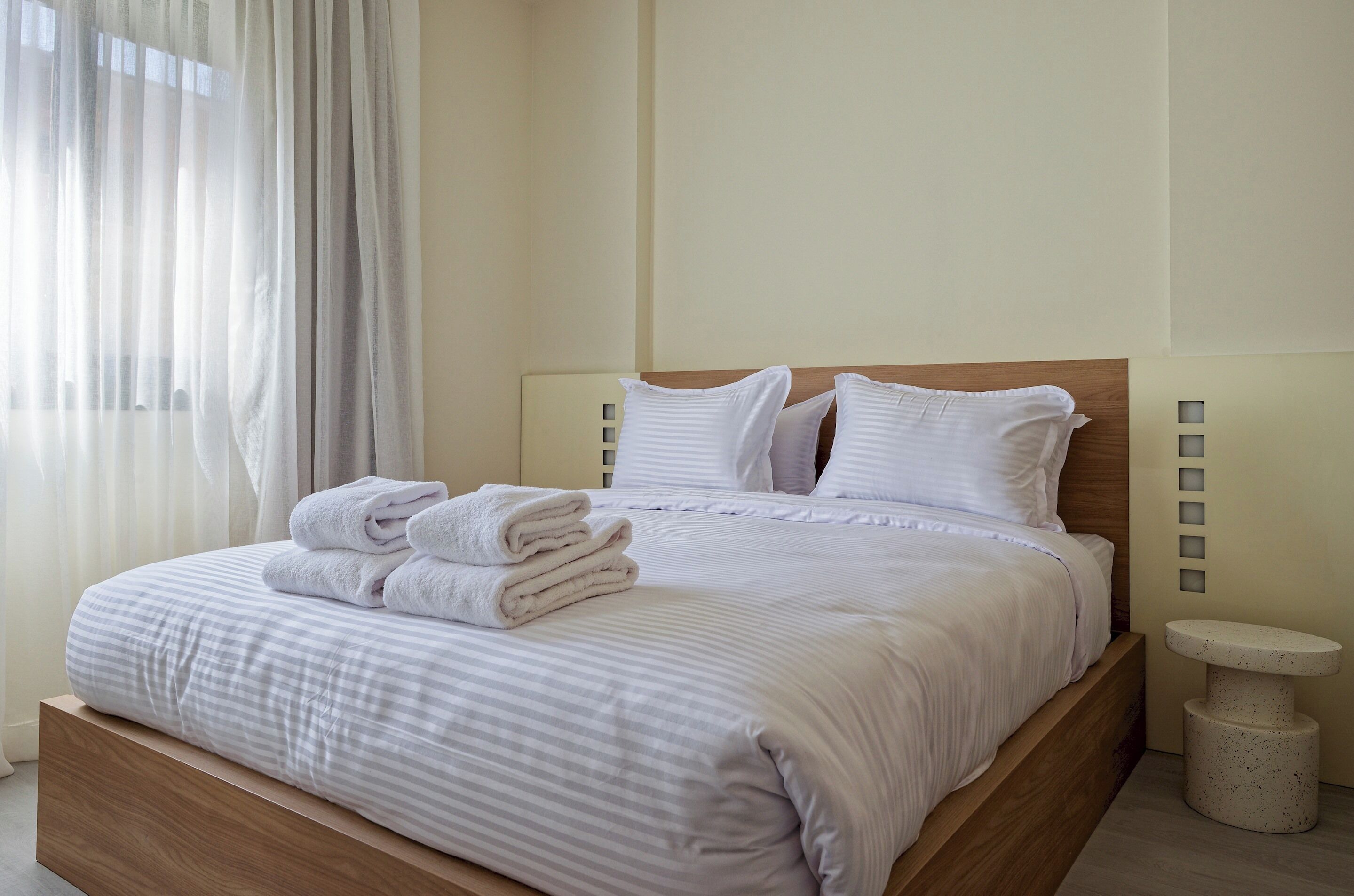 Un lit bien fait, avec du linge de lit et des serviettes blanches, une tête de lit en bois et une petite table ronde placée à côté.