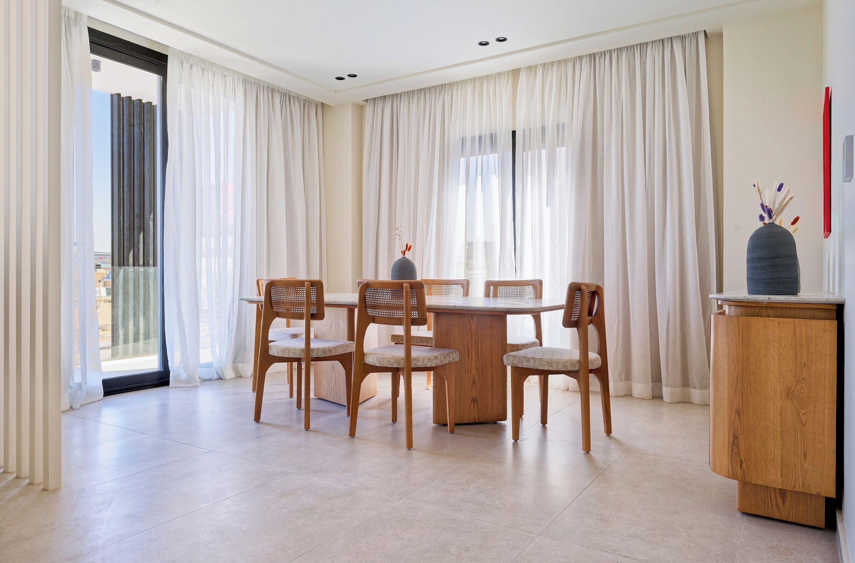 Uma sala de jantar com uma mesa redonda, quatro cadeiras de madeira, um armário e vista através de uma porta de vidro.