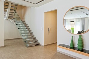 A modern interior with a glass staircase, a wooden door, and a round mirror on the wall. - VESTA  - Luxury Duplex - 3BR - Zayed City I (Sheikh Zayed City)