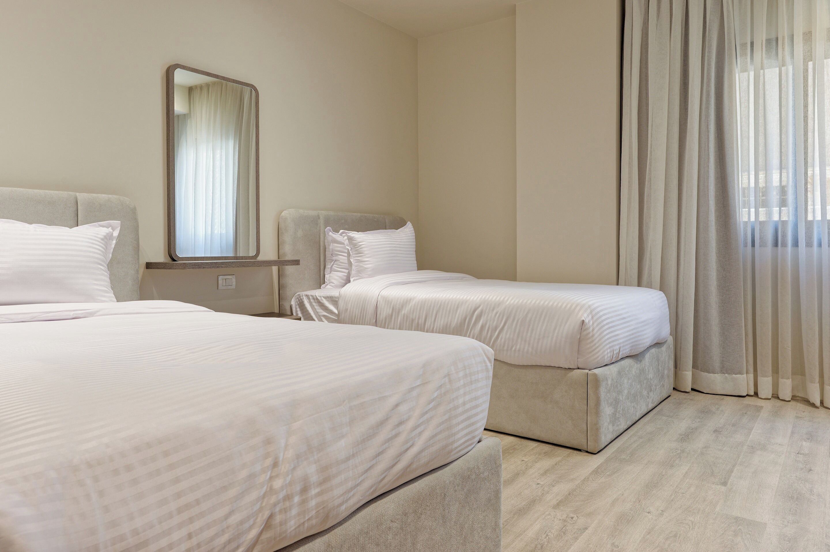 A hotel room with two beds, a large mirror, and sheer curtains.