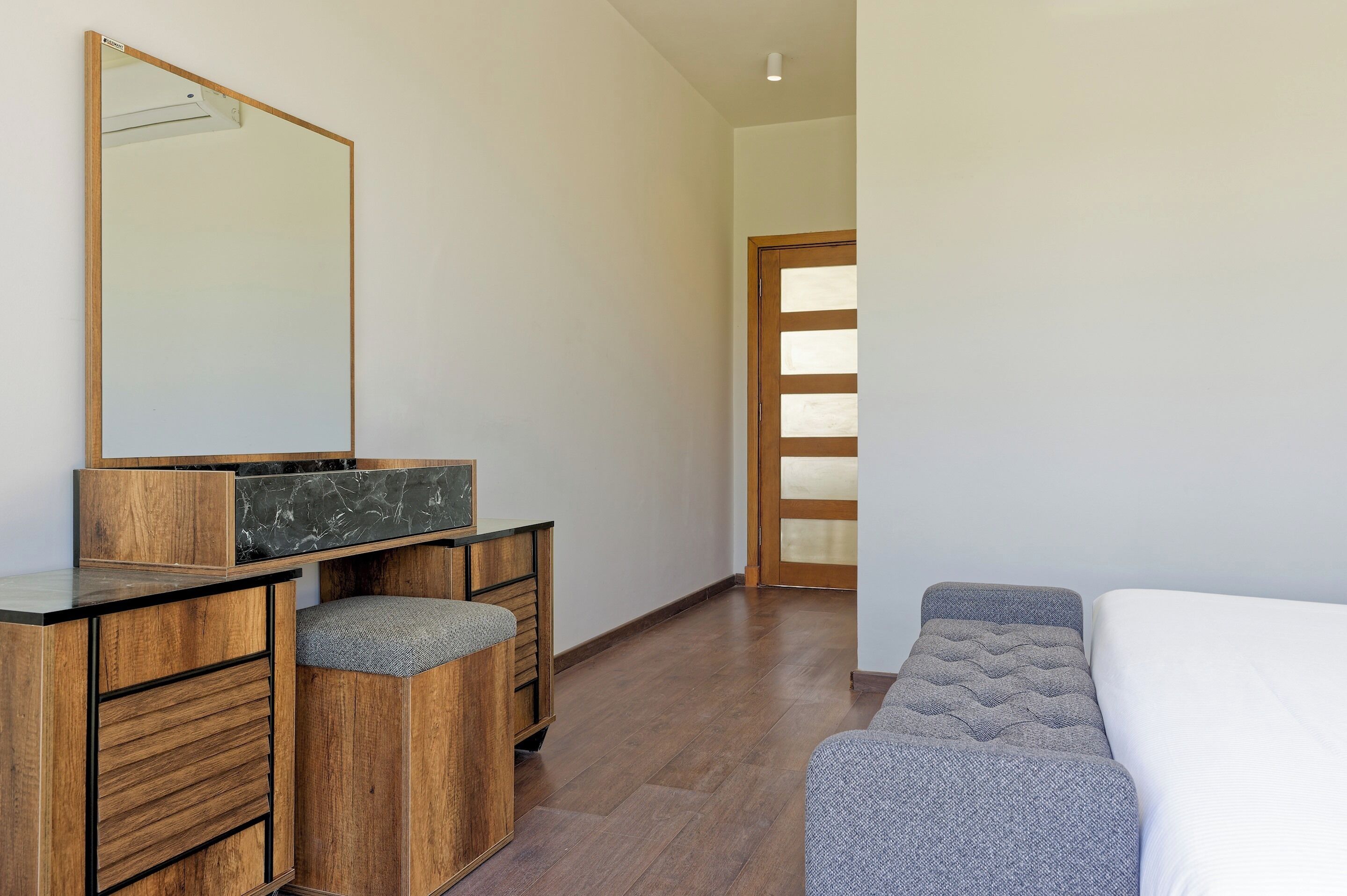 Une chambre avec une commode en bois, un grand miroir, un lit avec une tête de lit grise et une porte en bois dotée de panneaux en verre dépoli.