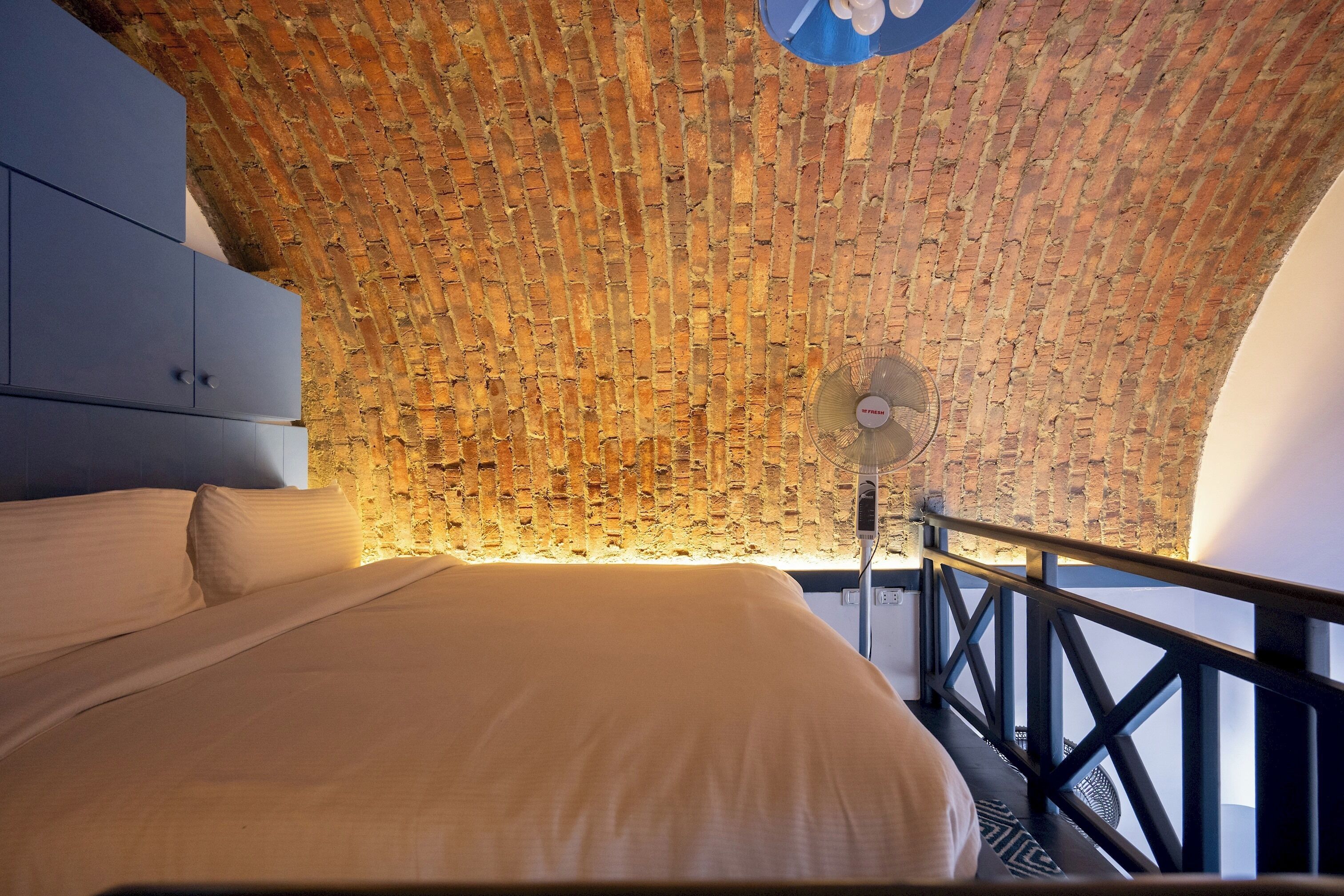 Une pièce avec un plafond en briques, un lit avec une literie blanche, une armoire bleue et un ventilateur sur pied.