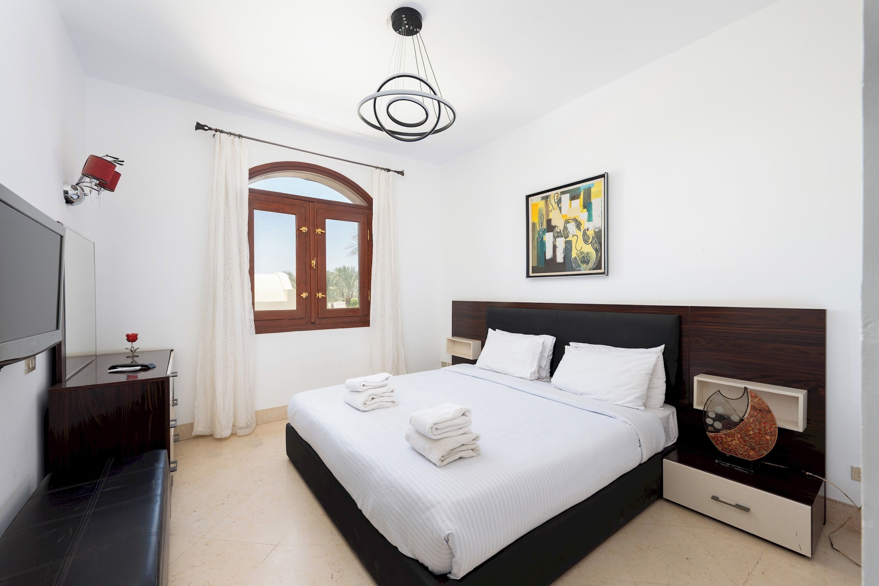 Una habitación de hotel con cama, televisión, escritorio, silla, ventana con cortinas y un cuadro en la pared.