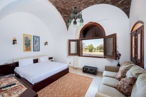 Une chambre avec un grand lit, un canapé, une vue sur l’extérieur par une fenêtre ouverte et un lustre.